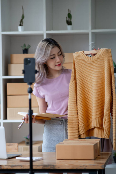 Young woman selling goods product clothes dress shirt online by smartphone live streaming video interaction internet online shop and talking to camera - Foto, afbeelding