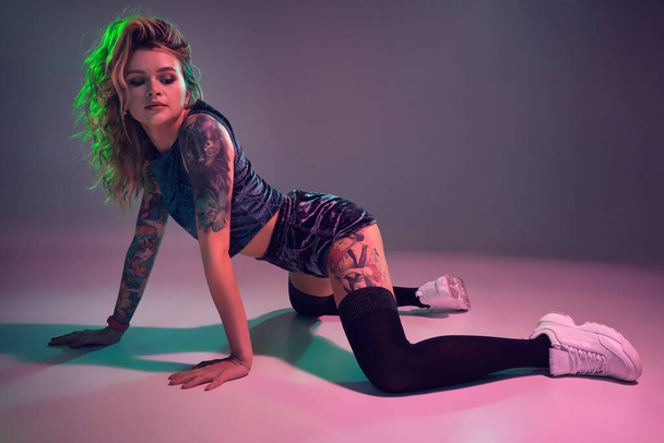 Joven hermosa chica rubia caucásica con tatuaje en su cuerpo, vistiendo pantalones cortos de terciopelo azul botín y medias negras bailando twerk en sus rodillas, fondo rosa
 - Foto, imagen