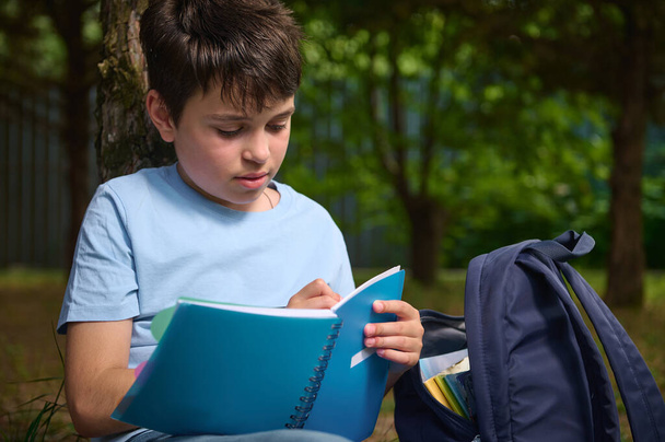 Close-up of a dark-haired Hispanic smart preteen school child, adorable teenage boy writing on copybook, doing homework outdoors. Back to school concept. Children. Lifestyle. Education concept - 写真・画像