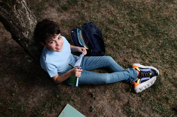 Vue aérienne d'un adolescent hispanique intelligent et plein de charme souriant en regardant la caméra, en écrivant, en prenant des notes sur le livre, en faisant des devoirs, en s'asseyant sur l'herbe dans le parc après les cours - Photo, image