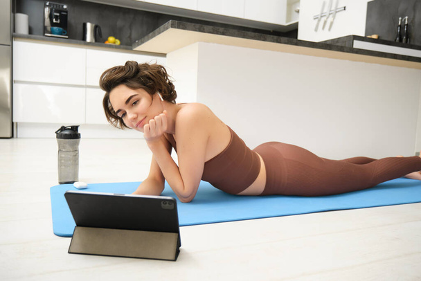 Jonge fitness vrouw, meisje in trainingspak liggend op fitness mat, kijken naar yoga, pilates video tutorial op tablet, doet workout thuis, maakt gebruik van gym app, training thuis. - Foto, afbeelding