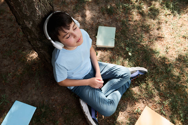 Widok na zrelaksowanego, przystojnego nastolatka słuchającego muzyki na słuchawkach bezprzewodowych, siedzącego na trawie w pobliżu szkolnych materiałów i kopiującego książki rozrzucone na trawie w ogródku lub parku - Zdjęcie, obraz