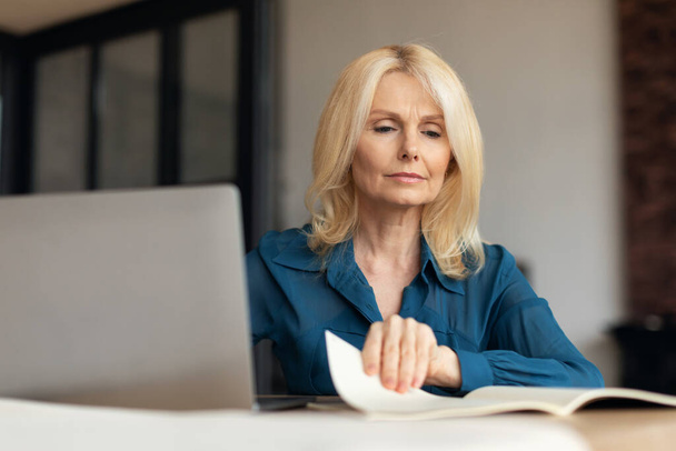 Ώριμη γυναίκα που εργάζεται με χαρτιά και laptop στο εσωτερικό του γραφείου, επιχειρηματίας που διαβάζει και ελέγχει έγγραφα, απολαμβάνοντας την απομακρυσμένη εργασία και τις επιχειρήσεις - Φωτογραφία, εικόνα