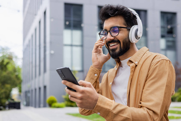 Young hispanic man in headphones uses an application for listening to online music and radio podcasts, a student smiles happily and types on the phone. - Fotoğraf, Görsel