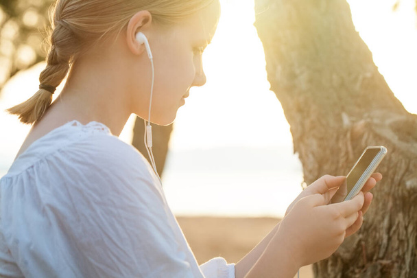 ヘッドフォンでブロンドの十代の少女の肖像画 携帯電話を保持し,海の背景に立っている - 写真・画像