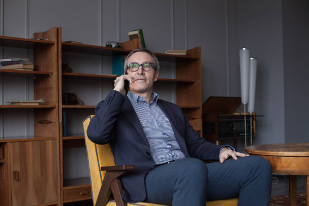 Homme d'affaires sérieux confiant avec les cheveux gris en costume et lunettes parler sur téléphone portable à l'intérieur sur fond de mur de bureau  - Photo, image