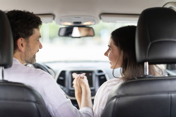 Back view of loving happy young couple sitting at front seats, holding hands and smiling to each other, handsome bearded man driving auto, taking his pretty girlfriend or wife on vacation, car trip - Foto, Imagen