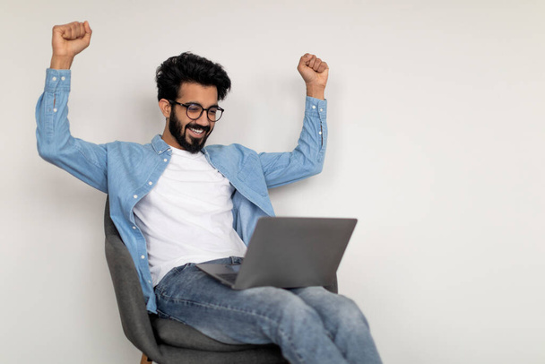 Онлайн-традинг. Счастливый молодой индийский парень, используя ноутбук и празднуя успех, переполненный Восточный человек дрожа кулаками, сидя на стуле над белой стеной фоне, приветствуя большую прибыль, скопировать пространство - Фото, изображение