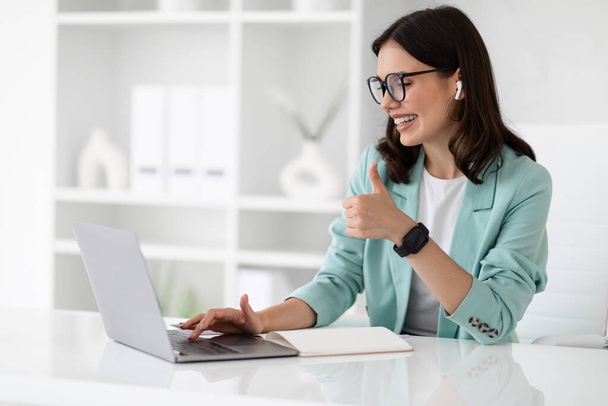 Glad young caucasian businesswoman in suit at table typing on computer, show thumb up in office interior. Meeting remotely, approve business, work and technology, communication - Photo, Image