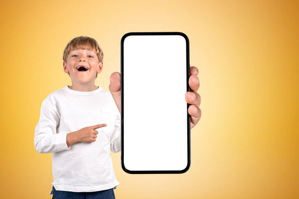 Excited school boy with opened mouth, finger pointing at large smartphone mock up screen, yellow background. Concept of social media and studies - Valokuva, kuva