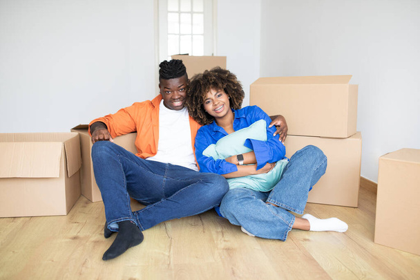 Έννοια ακινήτων. Ευτυχισμένο μαύρο ζευγάρι ποζάρουν στο πάτωμα στο νέο τους διαμέρισμα, χαρούμενα νεαρά αφρικανικά αμερικανικά συζύγους κάθεται ανάμεσα σε κουτιά χαρτόνι μετά τη μετακίνηση, χαρούμενη ιδιοκτήτες σπιτιού χαμογελώντας στην κάμερα - Φωτογραφία, εικόνα