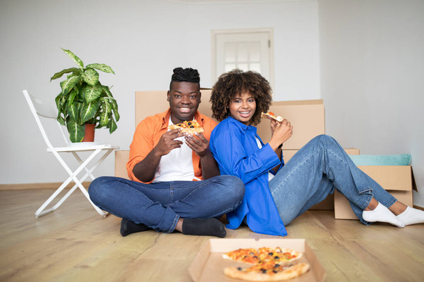 Mittagspause. Glückliche schwarze Ehepartner, die Pizza essen, während sie den Einzug in ihr neues Zuhause feiern, fröhliche junge afroamerikanische Paare, die es sich zwischen Pappkartons im Wohnzimmer gemütlich machen und in die Kamera lächeln - Foto, Bild