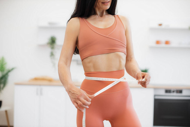 Cropped view of slender female holding measuring tape along waistline while standing in modern kitchen. Sporty brunette in terracotta activewear checking successful results of losing weight plan. - Valokuva, kuva