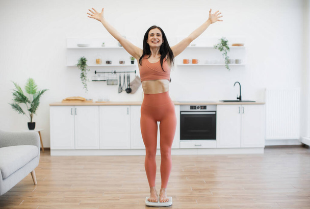 Full length view of woman in stylish activewear raising arms in victory gesture while standing on bath scales at home. Vigorous caucasian lady celebrating achievements in healthy lifestyle changes. - Foto, afbeelding