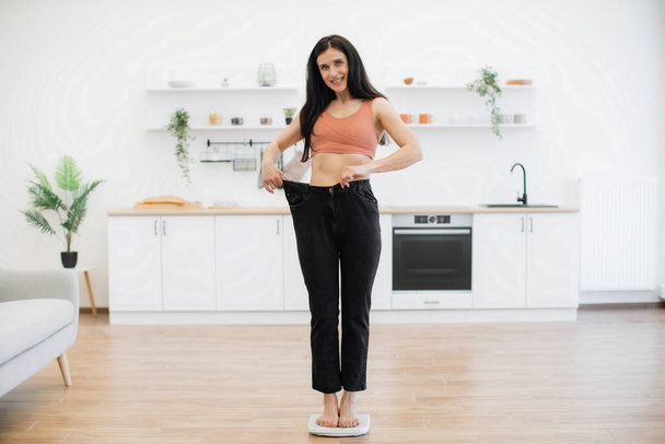 Amazed brunette female staring at loose pants on slim figure while standing on scales in middle of modern kitchen. Happy woman feeling pleasant shock of attained success in nutritional therapy. - Fotografie, Obrázek