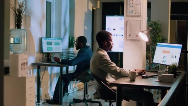 Retrato de un empleado afroamericano feliz trabajando de noche con su compañero de trabajo en la oficina. Colegas meticulosos en el espacio de trabajo que realizan tareas informáticas durante la noche para el proyecto de equipo antes de la fecha límite - Imágenes, Vídeo
