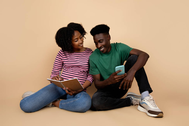 Positive young black man and lady with big belly sit on floor, enjoy pregnancy, make notes use smartphone app, isolated on beige background, studio. Parents, relationships and planning with device - Photo, Image