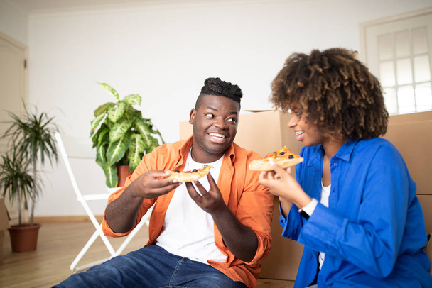 幸せな黒い配偶者はピザを食べて,移動日に楽しいを持っています, ボール紙箱の中で床に座っている陽気な若いアフリカ系アメリカ人のカップル, ランチブレイク, 休憩, 閉じるショット - 写真・画像