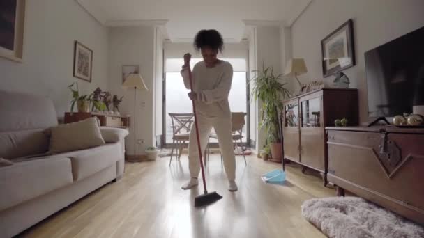 Genç Afrikalı Amerikalı kız süpürgeyle dans ediyor ve şarkı söylüyor. Ev işi oturma odası yapıyor. Siyah insanlar evi temizlemek için yerleri fırça ve faraşla süpürüyor. Neşeli kadın ev hayatı yapıyor.. - Video, Çekim