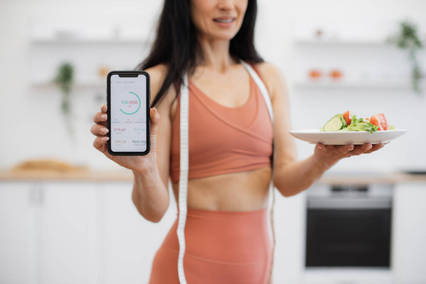 Crop of female person in sportswear posing with smartphone and fresh salad and showing amount of consumed calories on screen. Energetic woman using fitness tracker for making daily diet meal plan. - Фото, зображення
