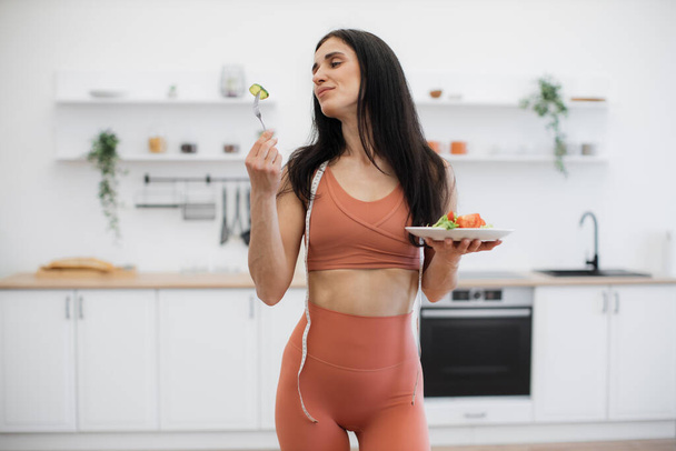 Portrait of slim beautiful fitness model posing with plate of nutritious snack and smiling at camera. Good-looking woman in tight sport clothing and measuring tape on neck leading healthy lifestyle. - Fotografie, Obrázek