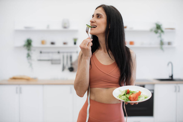 Long-haired brunette looking aside while trying freshly prepared vegan salad in spacious white kitchen on blurred background. Gorgeous woman promoting healthy eating for excellent well-being. - Fotó, kép