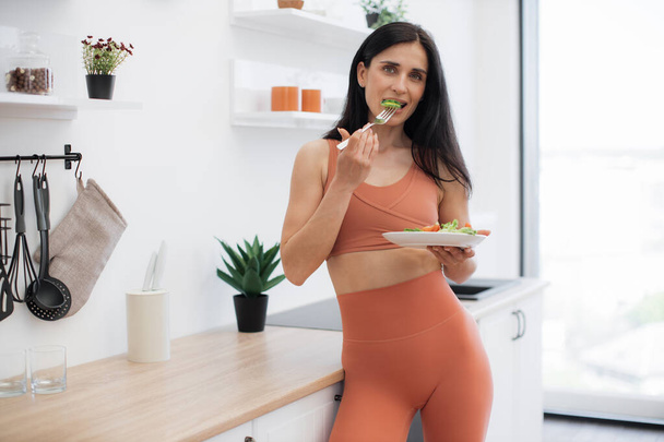 Pensive fitness person dressed in gym clothes standing in kitchen and looking at plate with greens. Long-haired brunette promoting healthy lifestyle with organic balanced food and regular training. - Valokuva, kuva