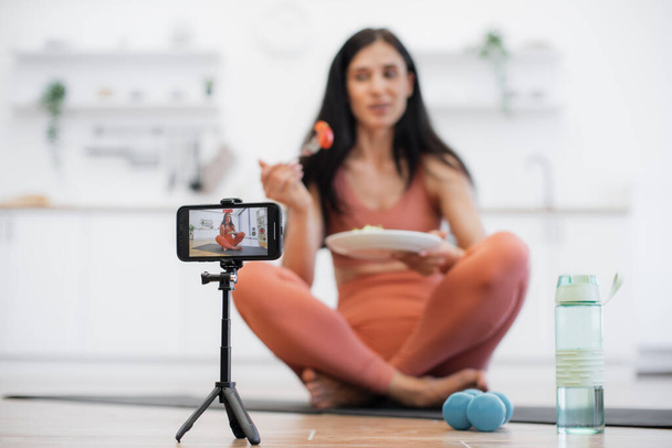 Blurred background of athletic woman posing on camera while sitting on rubber mat with plate full of organic meal. Fit lady filming video about healthy eating and physical exercising at home. - Zdjęcie, obraz