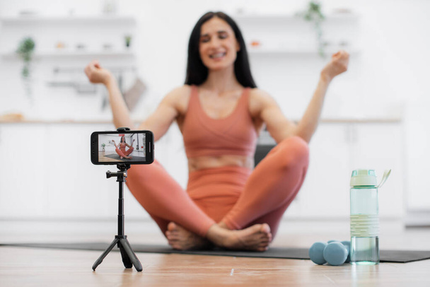 Professional female trainer recording video using tripod and smartphone during physical activity at home. Blurred background of woman meditating and smiling at camera while sitting in lotus pose. - Zdjęcie, obraz