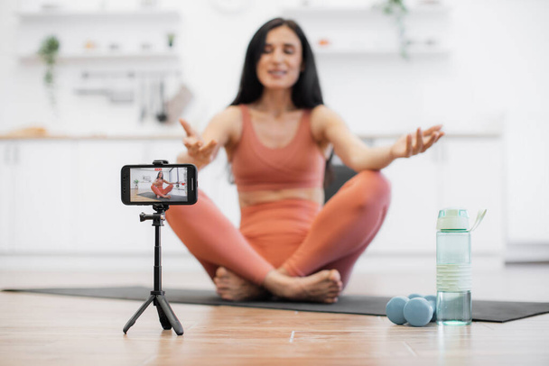 Professional female trainer recording video using tripod and smartphone during physical activity at home. Blurred background of woman meditating and smiling at camera while sitting in lotus pose. - Photo, Image