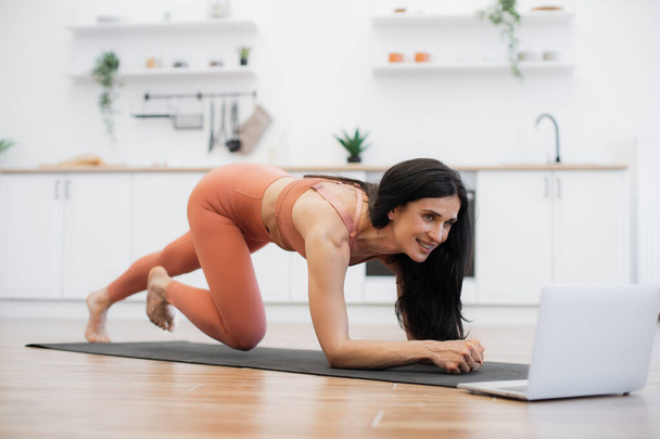 Athletic female with dark hair practicing on yoga mat in cozy kitchen and following online instructions of fitness coach on laptop. Skilled woman standing in plank position and bending right leg. - Foto, afbeelding