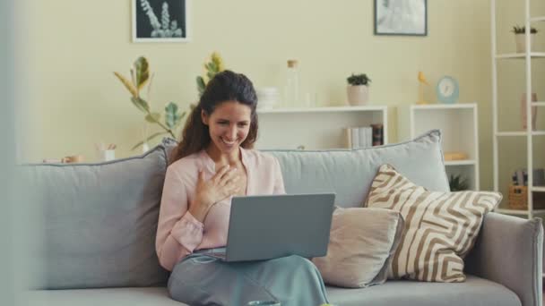 Όμορφη νεαρή μελαχρινή γυναίκα βίντεο κουβέντα με φίλο στο φορητό υπολογιστή, ενώ κάθεται στον καναπέ στο φωτεινό μοντέρνο σαλόνι - Πλάνα, βίντεο