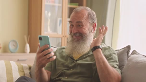 Bärtiger alter erwachsener Mann aus dem Kaukasus mit drahtlosen Ohrstöpseln, der tagsüber auf der Couch im Wohnzimmer sitzt und mit der Familie per Smartphone chattet - Filmmaterial, Video