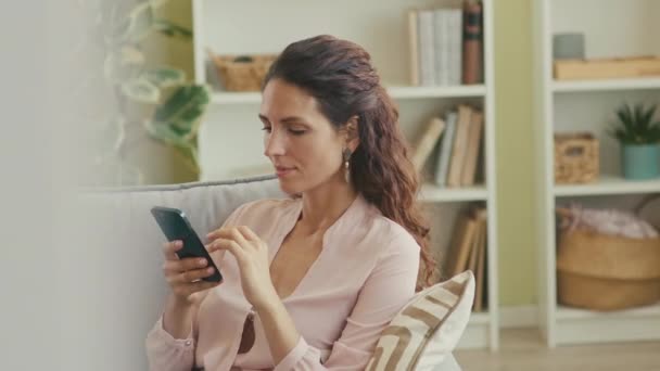 Young beautiful Caucasian woman with long brown hair scrolling on smartphone while sitting on soft couch in contemporary living room - Filmagem, Vídeo