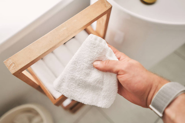 Ένας άντρας κρατάει μια λευκή πετσέτα. Μια στοίβα από λευκές βαμβακερές πετσέτες σε μια τουαλέτα. Στοίβα από λευκές πετσέτες terry είναι σε ξύλινο κουτί. Θραύσμα εσωτερικού του μπάνιου. Υψηλής ποιότητας φωτογραφία - Φωτογραφία, εικόνα