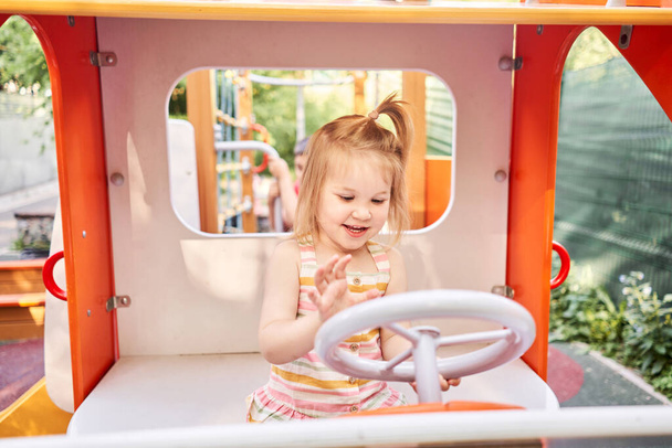 Little girl playing, she is a car driver holding steering wheel on playground. A sunny summer evening, a fun childhood. High quality photo - Photo, Image