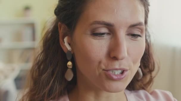Közelkép mosolygós fiatal kaukázusi barna nő vezeték nélküli fülhallgató beszél video chat otthonról - Felvétel, videó