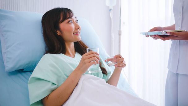 手術後にベッドに横たわるアジア女性に薬を与える女性看護師. - 写真・画像