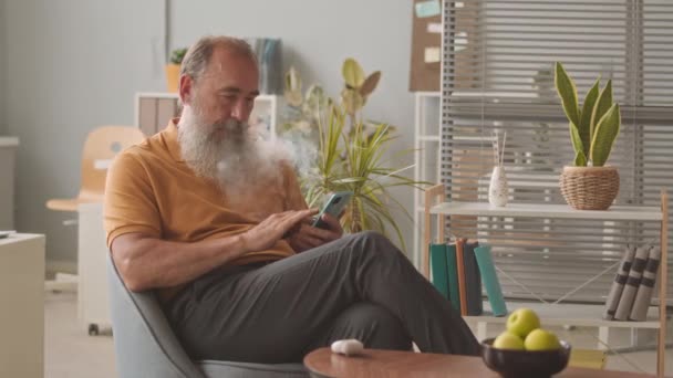 Brodaty starzec przewijanie wiadomości na smartfonie i palenie papierosa elektronicznego, siedzi na krześle w biurze - Materiał filmowy, wideo