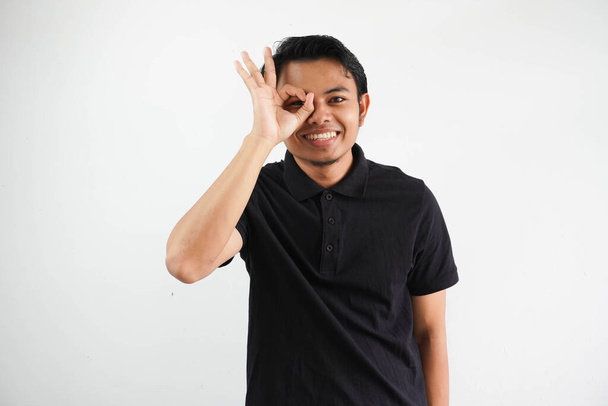 jovem asiático homem animado mantendo ok gesto no olho, vestindo preto polo t shirt isolado no branco fundo - Foto, Imagem