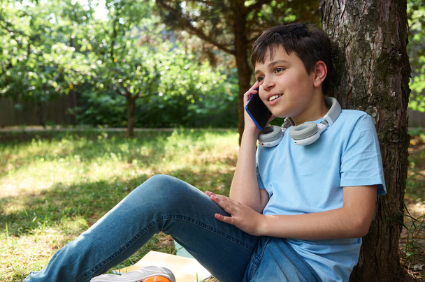 Retrato autêntico de um adolescente bonito feliz, estudante 11-13 anos de idade falando no telefone celular inteligente enquanto relaxa no parque depois da escola. Filhos. Pessoas pré-adolescentes. Estilo de vida. Lazer - Foto, Imagem