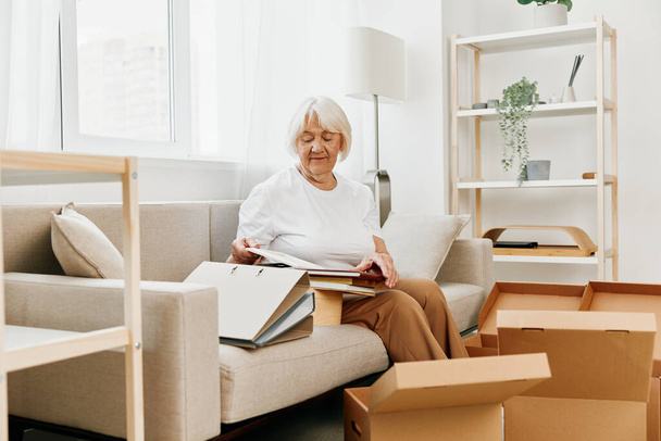 ηλικιωμένη γυναίκα κάθεται σε έναν καναπέ στο σπίτι με κουτιά. Συλλογή πραγμάτων με αναμνήσεις και συγκινητικό χαμόγελο και χαμόγελο ευτυχίας. Υψηλής ποιότητας φωτογραφία - Φωτογραφία, εικόνα