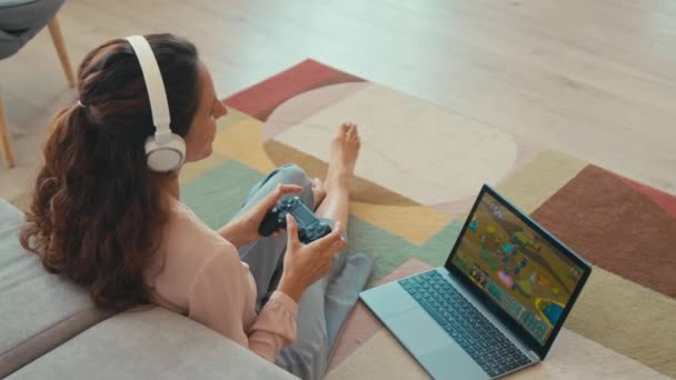 Молода жінка з довгим коричневим волоссям у бездротових навушниках виграє під час гри на ноутбуці з контролером, сидячи на килимі у вітальні - Кадри, відео
