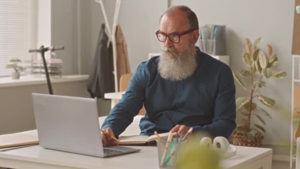 Közepes lövés az öreg fehér üzletember hosszú szürke szakállú laptoppal a munkahelyen - Felvétel, videó