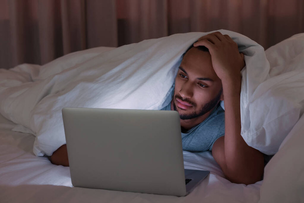 Young man using laptop under blanket in bed at night. Internet addiction - 写真・画像