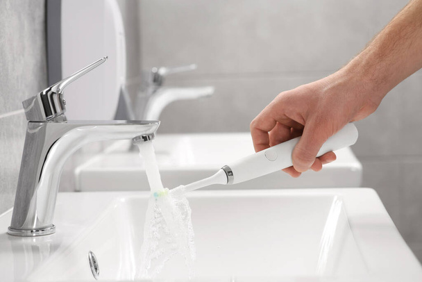 Man holding electric toothbrush under flowing water above sink in bathroom, closeup - Фото, изображение