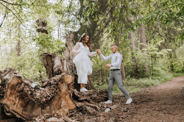 Esküvői séta az erdőben. A vőlegény fogja a menyasszony kezét, és egy nagy fatörzsön állnak. Függőleges fénykép - Fotó, kép