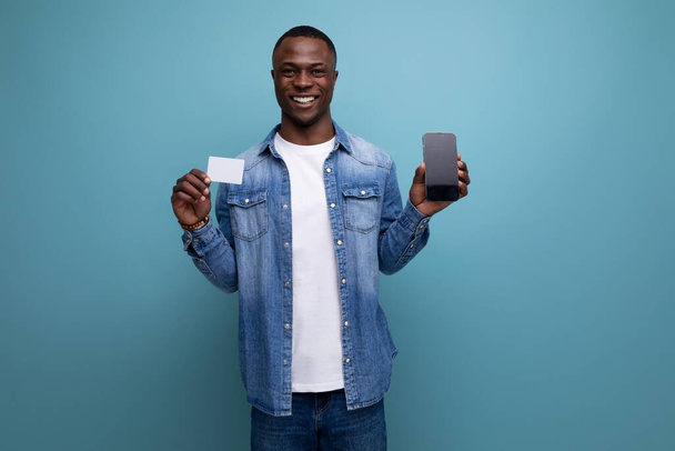 jeune beau jeune Africain en vêtements urbains tenant carte en plastique et smartphone maquette sur fond isolé avec espace de copie. - Photo, image