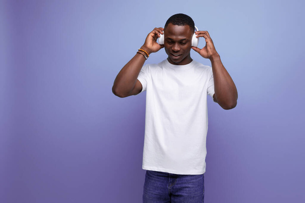 enérgico alegre chico americano de piel oscura en una camiseta blanca escucha un podcast en auriculares. - Foto, imagen