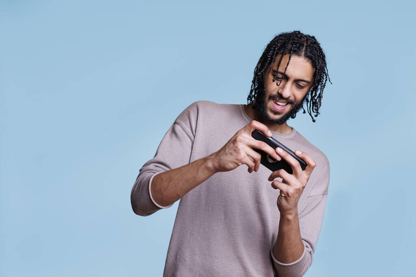 Lachende jonge Arabier man spelen video game op de smartphone en plezier hebben. Vrolijke persoon met behulp van onderhoudende mobiele telefoon software applicatie, genieten van online vrijetijdsbesteding - Foto, afbeelding